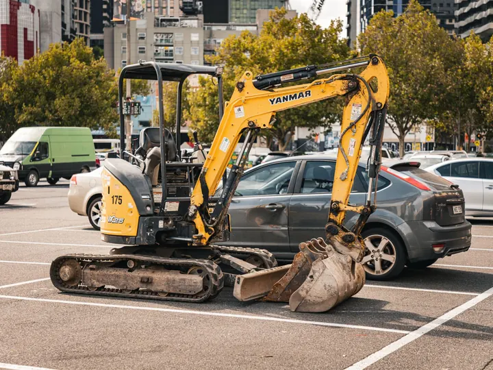 Parked mini excavator in a public parking lot in Melbourne, Australia, surrounded by cars and urban buildings.