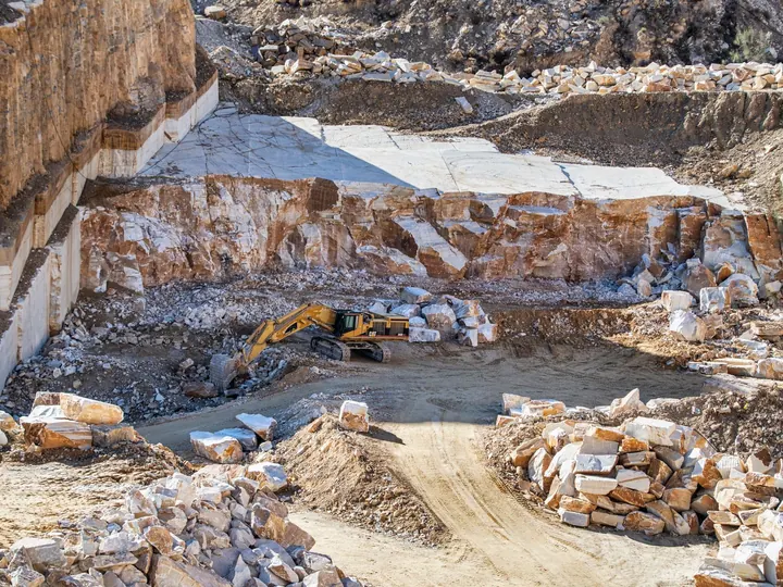 An excavator operates in a large stone quarry with rugged rocky terrain under daylight.