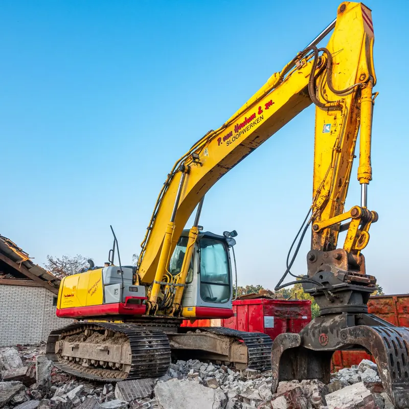 A large yellow excavator working amidst rubble at a construction site under a clear blue sky.