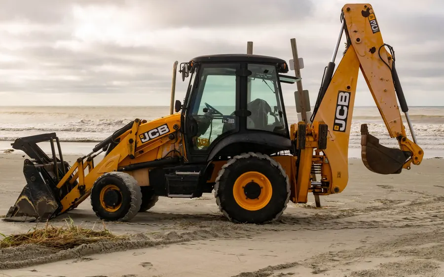 A JCB backhoe parked on the sandy shores of Sunset Beach, North Carolina with ocean waves in the background.