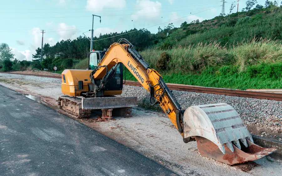 Yellow excavator parked on a construction site beside railroad tracks and greenery.
