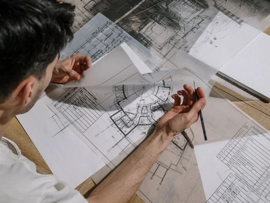 Architect studying and sketching over detailed architectural blueprints indoors.