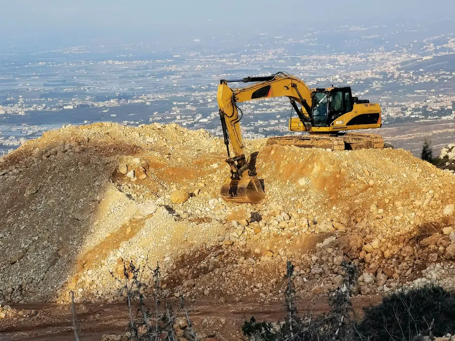 Yellow excavator on a rocky hilltop with a panoramic cityscape in the background, showcasing construction work.