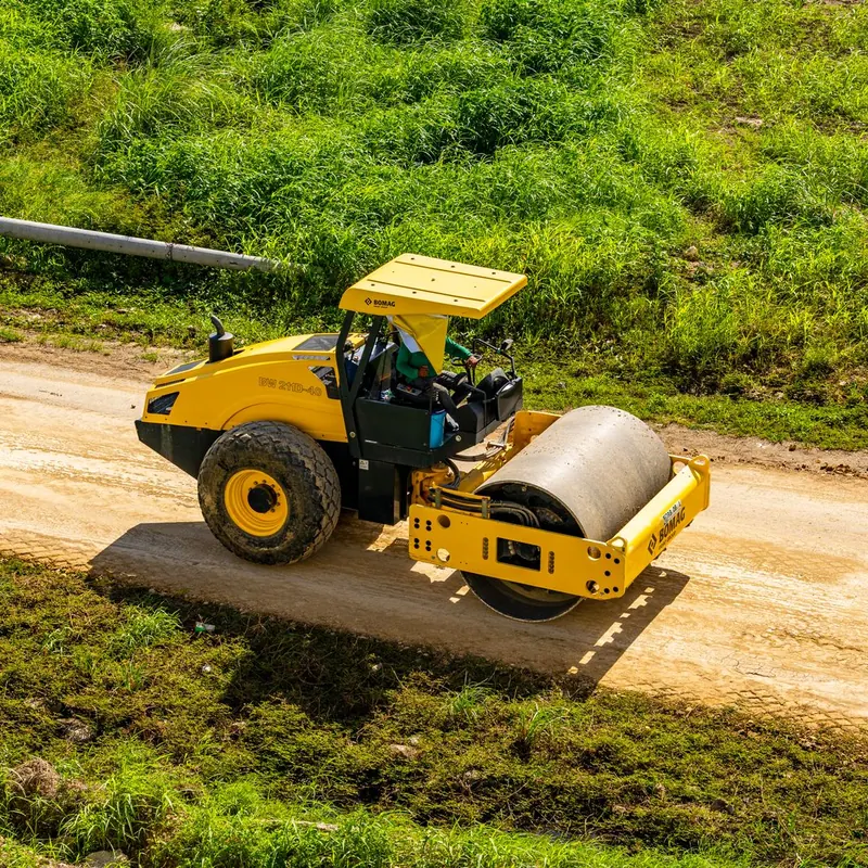 Aerial shot of a worker operating a road roller on a rural dirt path with greenery around.
