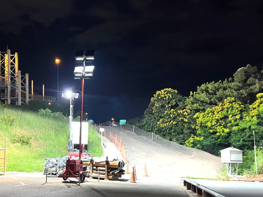 Torre de iluminação acesa em obra rodoviária à noite, com cones de sinalização